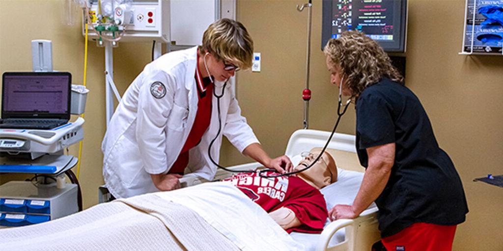 Two healthcare professionals examine a medical mannequin in a clinical setting, using stethoscopes and medical equipment to simulate patient care.